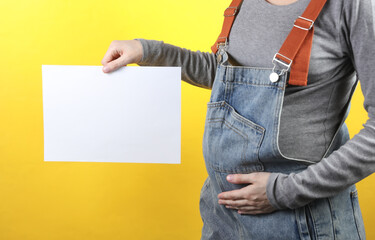 Pregnant woman in denim overalls holding blank paper sheet for your text on yellow background