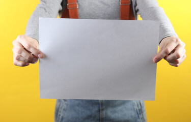 Pregnant woman in denim overalls holding blank paper sheet for your text on yellow background