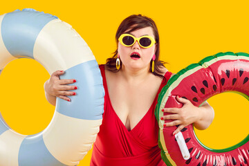 Overweight woman holding two inflatable swimming rings and looking at you in surprise and shock. Chubby woman in swimsuit and sunglasses on orange background. Summer holiday trip and vacation concept.