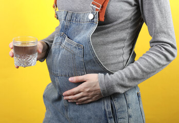 Pregnant woman in denim overalls holding glass with water on yellow background