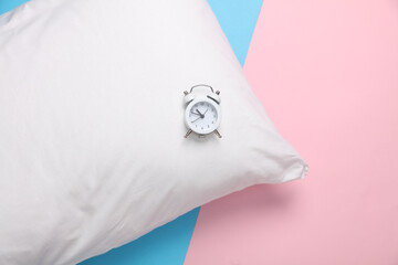White pillow with alarm clcok on a blue-pink pastel background. Time to sleep. Top view