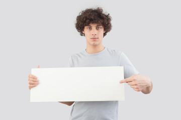 Handsome young man, pointing with finger to show a blank white signboard, isolated on gray background. Placard copy space for text or logo, banner for advertising or promotional messages