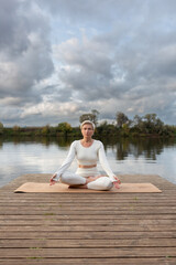 A beautiful adult woman with short hair is sitting on the pier in the lotus position, doing yoga