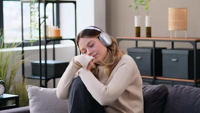 Discontent young Caucasian woman in headphones sitting on sofa, listening music at home. Sadness, hopelessness, boredom, apathy concept.