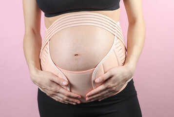 Young Pregnant woman with orthopedic belt for back and belly support. Bandage for pregnant women. Special belt for the 3rd trimester. Studio shot on pink background