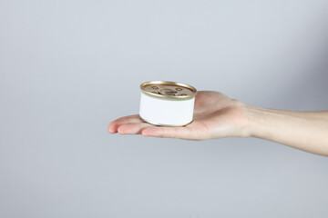 Tin can of canned food with a white label, mockup for your design in a female hand on a gray studio...