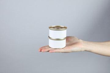 Tin cans of canned food with a white label, mockup for your design in a female hand on a gray...