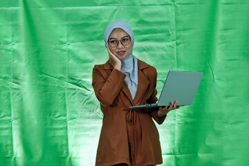 cheerful young Asian woman wearing hijab, glases and blazer holding laptop and thinking about...