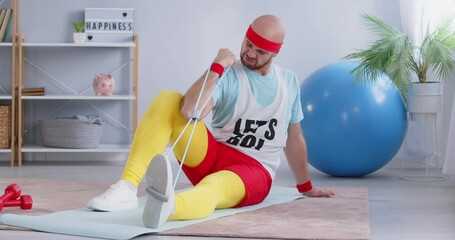 Fun and unconventional approach to fitness. Funny man in in humorous retro clothes using resistance band as part of his workout routine. Man is exercising while sitting on sports mat at home. Footage.