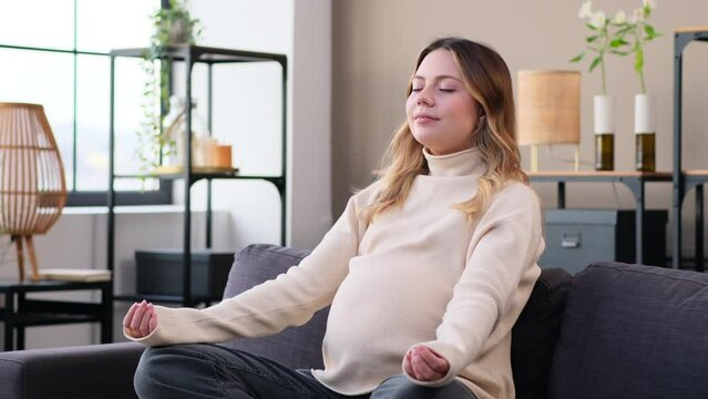 Friendly pregnant Caucasian woman meditating on sofa at home living room. Yoga practice, future maternity, domestic relaxing concept.