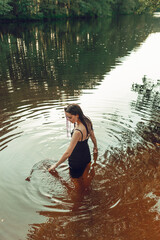 Calm woman in wet dress standing in lake and touching water in summer forest