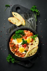  Tasty Breakfast Shakshuka in an Iron Pan. Fried eggs with tomatoes. On a black stone background. © Yaruniv-Studio