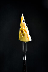 A triangle of Maasdam cheese with large holes on a metal fork. On a black background, vertical photo. - 788118416