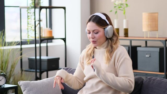 Caucasian cheerful young woman listening music using headphones and dancing on sofa at home living room. Celebrating weekend relaxation.