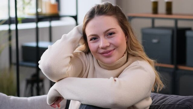 Portrait of a smiling and positive Caucasian young woman, relaxing on comfortable sofa at home living room. Content and satisfied lady looking at camera.