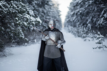 Naklejka premium Portrait of a medieval fantasy warrior in a horned helmet, steel cuirass, chain mail with a two-handed ax in his hands, standing in a fighting position against the backdrop of a winter forest.
