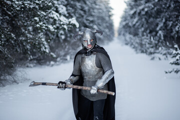 Portrait of a medieval fantasy warrior in a horned helmet, steel cuirass, chain mail with a two-handed ax in his hands, standing in a fighting position against the backdrop of a winter forest. - obrazy, fototapety, plakaty