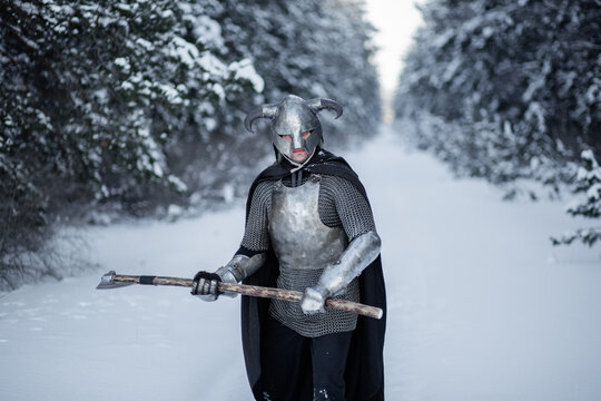 Portrait of a medieval fantasy warrior in a horned helmet, steel cuirass, chain mail with a two-handed ax in his hands, standing in a fighting position against the backdrop of a winter forest.