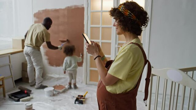 Side footage of expecting Caucasian mother texting on phone and searching for design ideas for future nursery or playroom while her husband painting walls brown with little daughter
