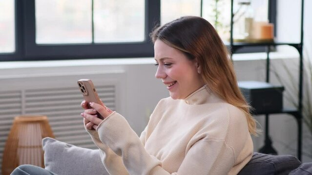 Smiling Caucasian woman messaging in social media or surfing internet and laughing using phone at home living room. Resting online.