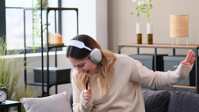 Cheerful Caucasian woman listening music in wireless headphones, dancing and singing song sitting on sofa at home. Spending leisure time, enjoying weekend.