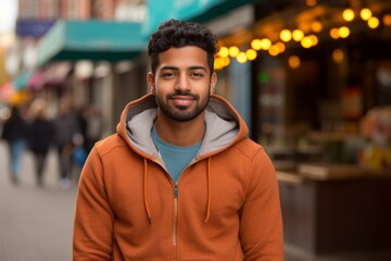 Fototapeta premium Portrait of a satisfied indian man in his 20s sporting a comfortable hoodie on vibrant market street background