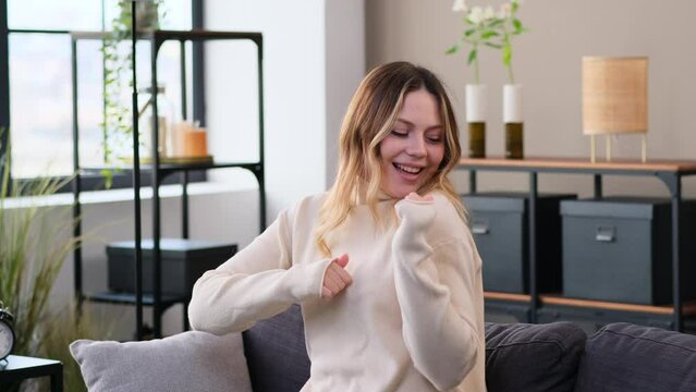 Happy Caucasian woman listening music and dancing on sofa at home. Celebrating weekend, enjoying relaxation and domestic entertainment.