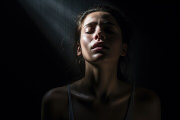 Young woman in dark room with light shining on her face, eyes closed and expression of pain or stress. Dramatic lighting creating shadow over head and body, highlighting facial features and emotions.