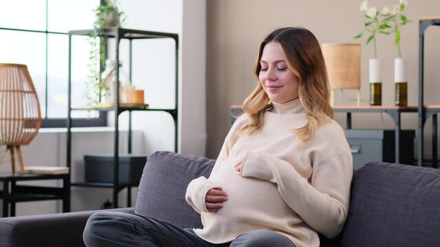 Happy Caucasian young pregnant woman touching belly on sofa at home living room. Resting at home, expecting childbirth and maternity.