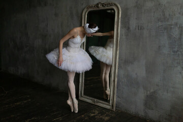 Ballerina in a swan costume stands on pointe shoes and looks in the mirror