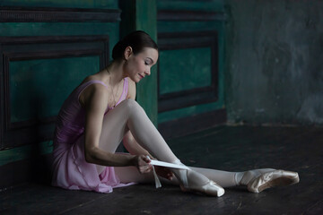 Ballerina in a pink dress puts on pointe shoes while sitting on the floor.