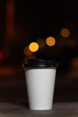 Close-up of a disposable white paper cup with coffee or tea stands on a bench against the background of a blurred city at night with lights. Coffee to go. Vertical photo
