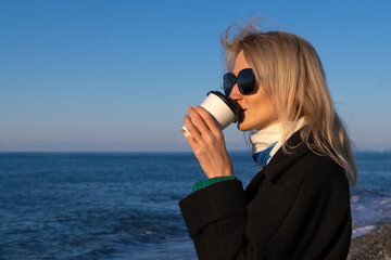 Young beautiful happy blonde woman in a black coat and sunglasses drinks takeaway coffee from a paper cup by the sea on a sunny day