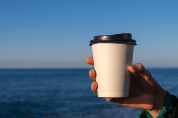Close-up of a woman's hand holding a white paper disposable cup with coffee or tea against the background of the sea on a sunny day, copy space