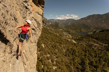 A woman is climbing a rock face with a red tank top on. The mountain range in the background is...