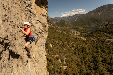 A woman is climbing a rock wall while wearing a red shirt and blue shorts. The scene is set in a mountainous area with a beautiful view of the surrounding landscape