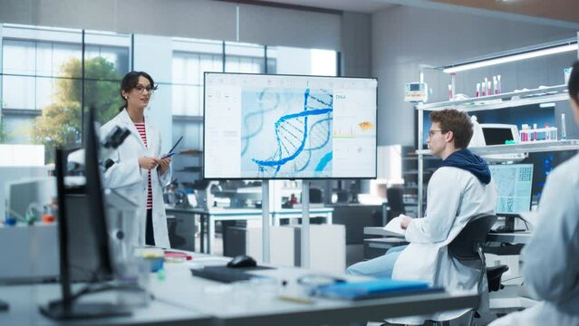 Group of Talented Female and Male STEM Students Having a Meeting with Their Project Leader in a Modern Laboratory. Young Female Specialist Showing a Presentation About Biochemistry on Screen