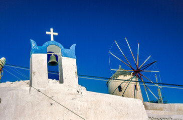 Glockenstuhl einer Kapelle und Windmühle in Unteransicht vor blauem, wolkenlosem Himmel auf der griechischen Insel San Turin im hellen Licht der Mittagssonne
