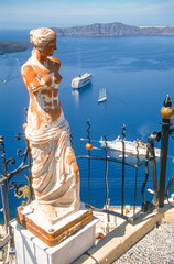 Eine Statue der griechischen Göttin Aphrodite am Kraterrand der Insel San Turin, mit Blick über den Abgrund und vor Anker liegenden Segel- und Kreuzfahrtschiffen - 788159250