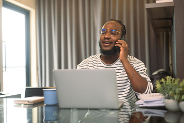 Smiling young African man in eyeglasses talking on mobile phone and using laptop at home