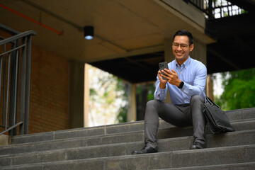 Full body shot of smiling Asian businessman sitting on stairs of office building and using smartphone