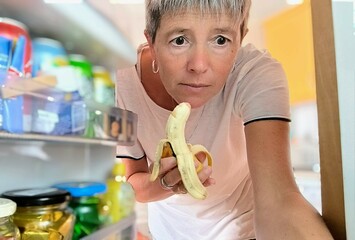 Woman seen from the refrigerator, holding a banana, with a fixed gaze looking inside.  Eating...