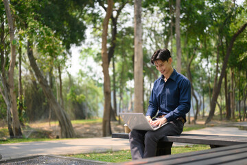 Smiling young businessman sitting on bench and using a laptop at public park