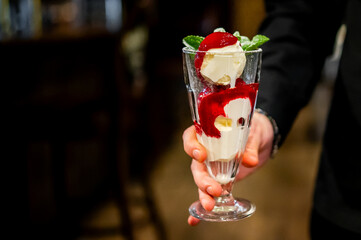 Person holding a glass dessert garnished with red syrup, cream, and fresh mint, capturing the indulgence of a beautifully presented treat.