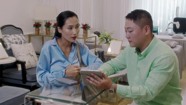 Medium shot of Asian male consultant showing list of available spa salon services on digital tablet to female client