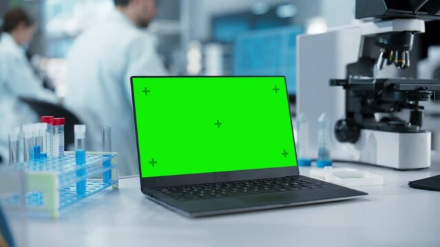 Laptop Computer with Green Screen Mock Up Display Standing on a Table in a Science Laboratory Next to a Microscope and Test Tubes with Biological Samples. Anonymous People Working in the Background