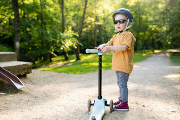 Adorable little boy riding his scooter in a city park on sunny summer evening. Young child riding a...