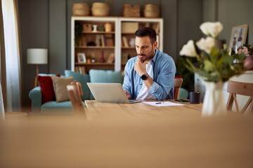 A focused male web designer resting his chin on his hand and sitting in front of his laptop going over his work.