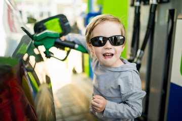Cute little blond boy holding pump nozzle. Small kid helping father to fuel the car at a gas station. Daddy's little helper. - obrazy, fototapety, plakaty
