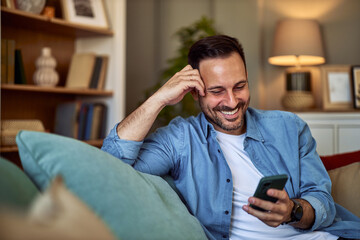 A happy young adult man reading a text message from his date while sitting and waiting on a sofa at...
